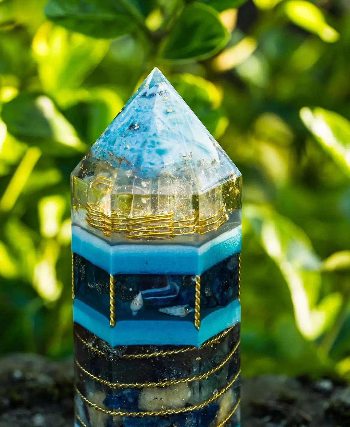 Orgonit-Turm mit blauen Schichten "Ein handgefertigter Orgonit-Turm mit pyramidenförmiger Spitze, blauen und transparenten Schichten, goldenen Spiralen und eingearbeiteten Edelsteinen. Die Struktur ist kunstvoll gestaltet und reflektiert das Sonnenlicht, während im Hintergrund grüne Blätter leuchten."