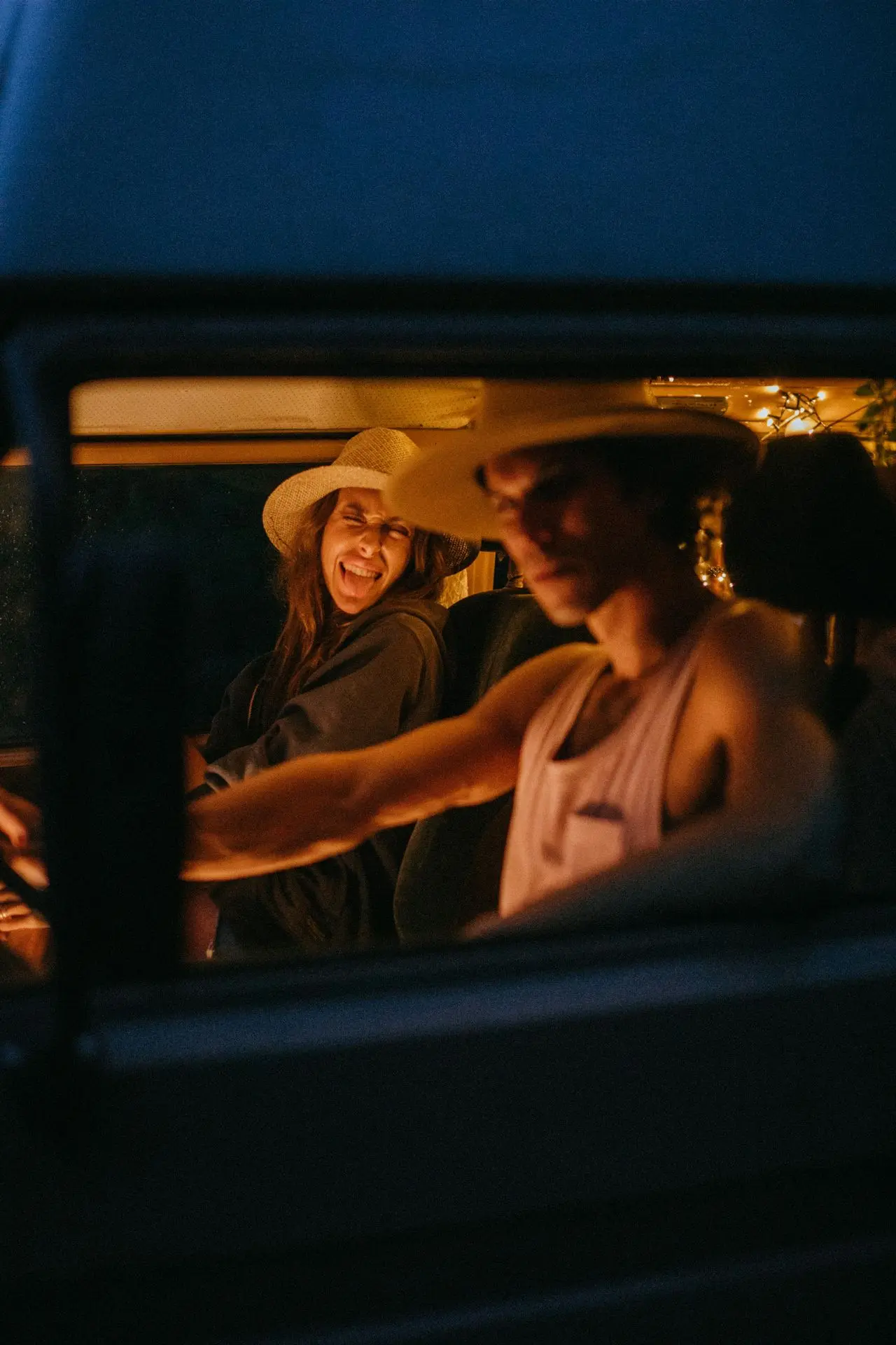 Roadtrip mit Spaßfaktor "Ein junges Paar in einem Van mit Strohhüten. Die Frau lacht und streckt spielerisch die Zunge raus, während der Mann konzentriert fährt. Die Lichter im Auto sorgen für eine gemütliche Atmosphäre."