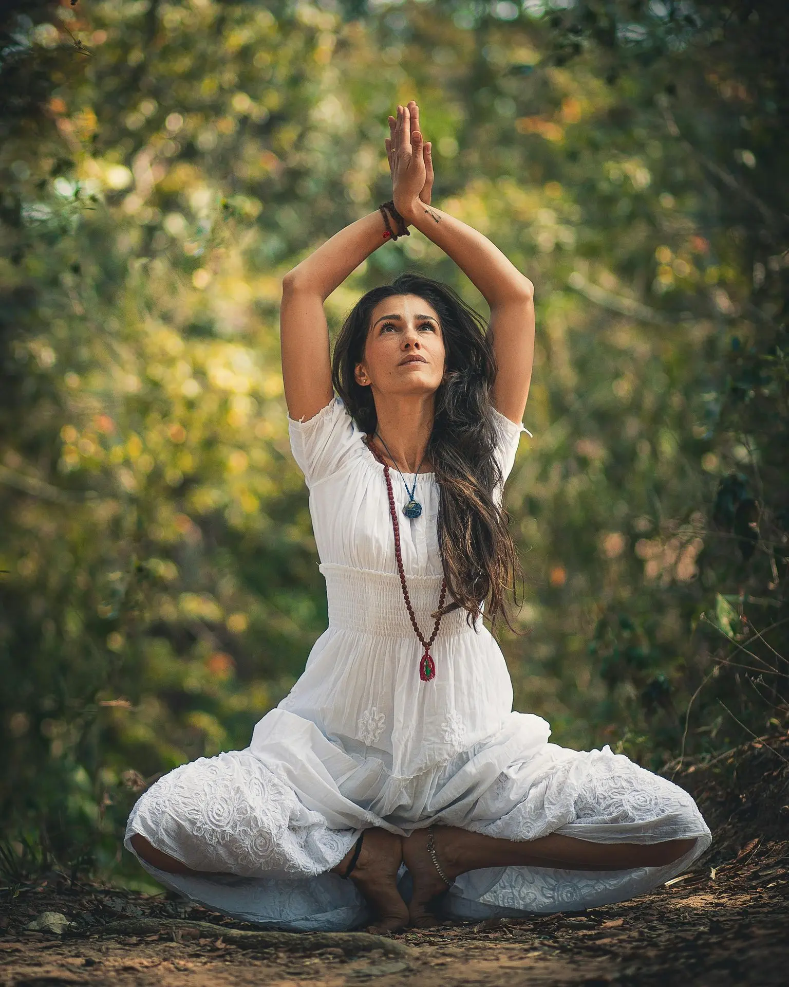 Spirituelle Yoga-Praxis im Wald "Eine Frau in weißem, fließendem Kleid sitzt meditierend im Wald mit erhobenen Händen. Sie trägt eine Kette mit Kristallen und strahlt innere Ruhe aus."