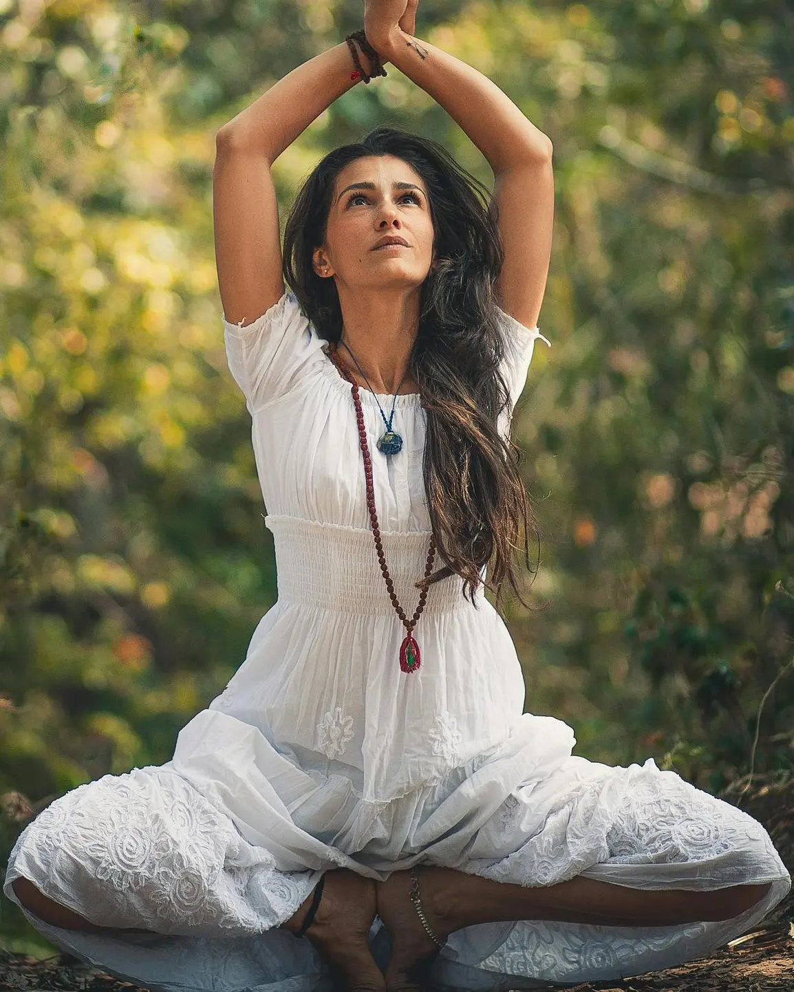 Spirituelle Yoga-Praxis im Wald "Eine Frau in weißem, fließendem Kleid sitzt meditierend im Wald mit erhobenen Händen. Sie trägt eine Kette mit Kristallen und strahlt innere Ruhe aus."
