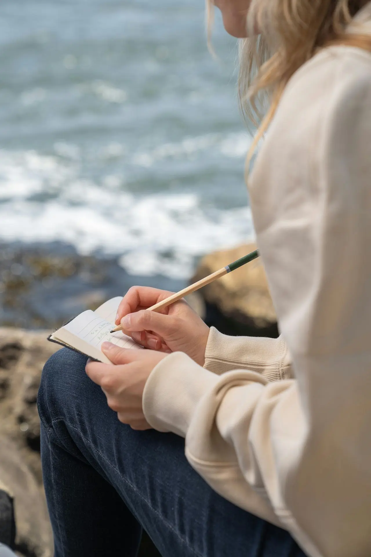 Tagebuchschreiben am Meer "Eine Frau in beiger Jacke sitzt auf einem Felsen am Meer und schreibt in ihr Notizbuch. Ihre blonden Haare wehen leicht im Wind."